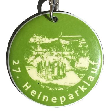 Grüner Medaillon mit der Aufschrift "27. Heineparklauf" und einer Parkszene.